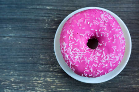Top vie to bright pink donut on white round plate and aged wooden grey background. Isolated object close up. Traditional American sweets for breakfast. Copy space. White sugar decoration on top.の写真素材