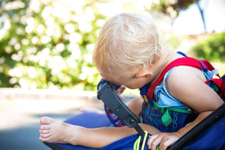 Blond kid sleeps in baby stroller with had on hand hold. Hot sunny summer day. Daytime sleep of child. Travelling with small children concept. Tired of heat. child safety equipmentの写真素材