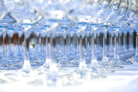 A lot of clean crystal glasses on white tablecloth prepared for big party. Blurred. Catering and restaurant utensils. wedding organisation. Wine tasting. Background picture. Group of objectsの写真素材