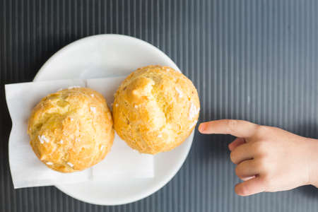 Child points with index finger two round eclair. Children and sweets context. Fresh italian cakes on white plate on grey surface. I want this. Bakery and kids. Gluten food for infantsの写真素材