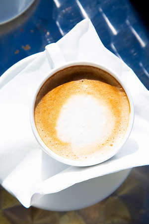 Top view to a hot fresh cup of capuccino in bright sunlight. White mug of hot coffee with milk on white napkin. Traditional Italian breakfast beverage. Vacation in Europe drink. Close upの写真素材