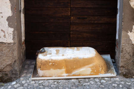 Old metal bath lyes upside down on cobblestone pavement outdoors in front of big wooden gate. Disposal of large-sized garbage. Vintage composition. Copy space. No people. Old city. Entrance blockedの写真素材