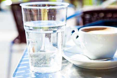 Transparent plastic glass with water with cup of cappuccino and caffe tables in background blurred. Traditional italian style of serving coffee. Still drinking water vs caffeine. Healthy dieting.の写真素材