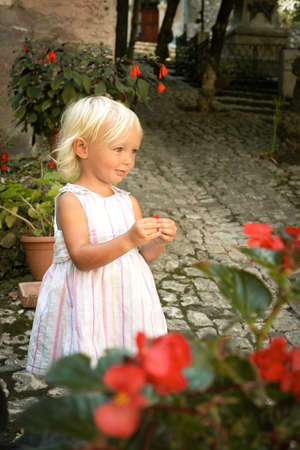 Beautiful blond blu eye caucasian baby girl in light summer dress with red flowers. Summer dreams concept. Childhood and travelling. Kid in old town. Positive emotionsの写真素材