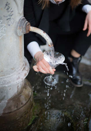 Traditional roman drinking fountain. Woman holds Martini cup. water falls in a crystal glass. Background blurred. Girl fills a glass with water. Drinking water. Stream. Pure water. Freshness concept.の写真素材