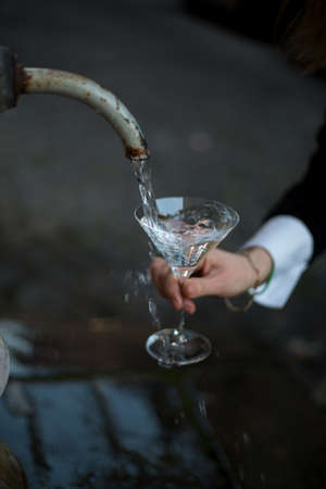 Martini crystal glass. Traditional roman street drinking fountain. Fresh water of antic aqueduct. Girl holds glass. office black and white suite. Background blurred. Drinking water. Freshness concept.の写真素材