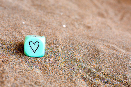 Light blu cube with heart and weather symbols on sides. Funny gambling dice on sand. Saint...