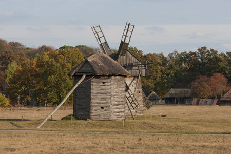 Autumnal country landscape. Two wooden windmills  in the foreground. Trees and small hut in the...