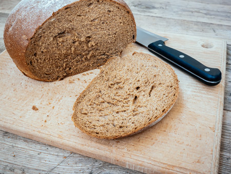 A slice of freshly baked wheat-rye bread with a crispy crust on a beech cutting board with a loaf...
