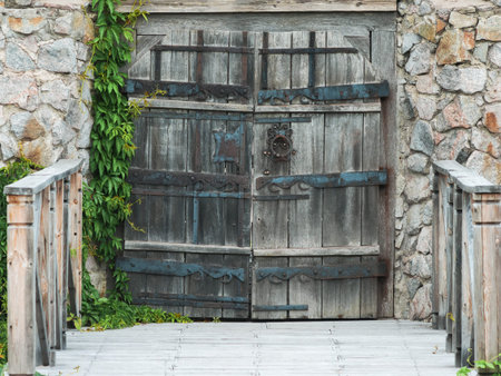 Old wooden gate with forged hinges in a stone wall overgrown with ivy and a bridge with wooden railings in front of itの写真素材