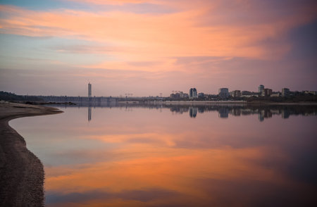 Cityscape during sunset on the bank of a wide river with a cable-stayed bridge and tall buildings on...