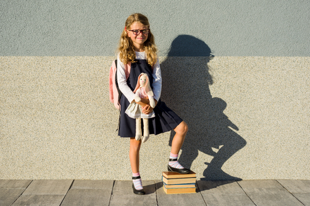 A cute schoolgirl of primary school is smiling with a toy in her hand. Girl with a backpack near the building outdoors. Leaving childhood 
and  Back to school!の写真素材