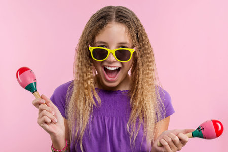 Cheerful teen girl with long blond curly hair. In bright yellow glasses she dances with maracas on a...
