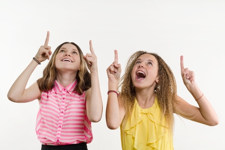 Attractive positive two teenage girl pointing her index finger up, indicating copy space on white blank wall for your content or information informationの写真素材