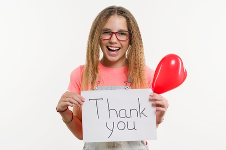 A teenage girl holding a paper with a thank you text, and a heart balloon, on a white studio backgroundの写真素材
