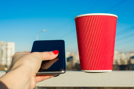 Red paper glass, phojne in a female hand, background urban, blue clear skyの写真素材