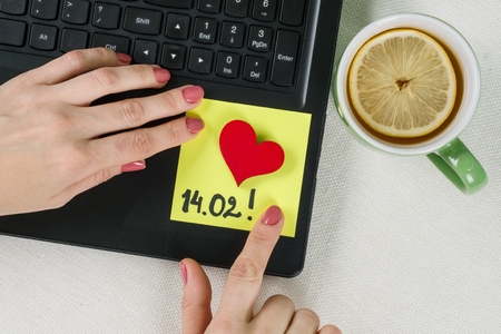 Valentine's Day. A note of text 14.02 written on a paper sticker. Background computer, laptop, woman's hands on the keyboard.の写真素材