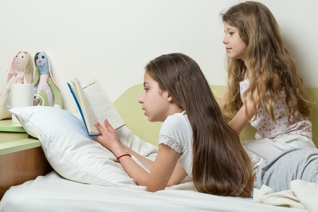 Children are reading a book lying in bed at home. The elder girl reads aloud to the younger sister.の写真素材