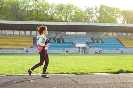 Portrait of middle aged woman running with headphones, bottle of water in the stadium in evening. Copy spaceの写真素材
