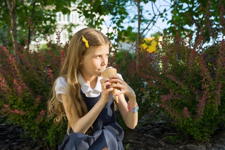 Schoolgirl 8 years old eating ice cream. Blond girl in school uniform, background of green trees, yard.の写真素材