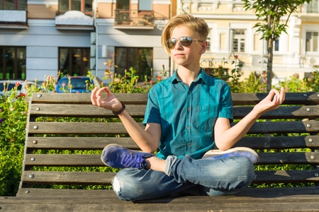 Yoga in the city, teenage boy sits in lotus pose on bench in city park. Relax, rest.の写真素材