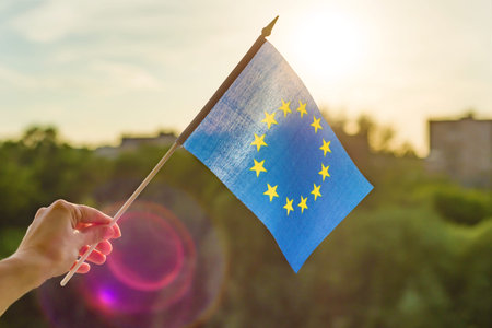 Hand holds European Union flag in an open window. Background blue sky, silhouette of the city,...