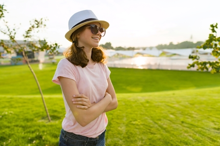 Outdoor portrait of young girl in sunglasses and hat with arms crossed. Background of green grass, recreation area, summer sunset. Copy spaceの写真素材