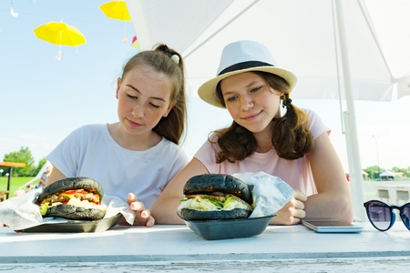 Adolescent girls with large black burgers. Sit in a street cafe, sunny summer day in recreation and entertainment areaの写真素材