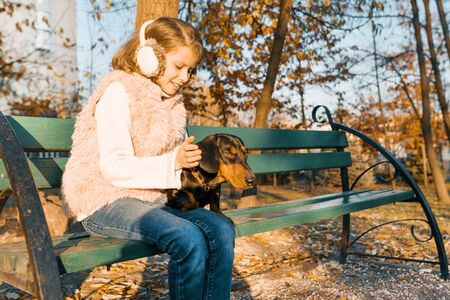 Smiling little owner of a dachshund dog sitting on bench in the autumn park, girl with love hugging dog at the golden hour.の写真素材