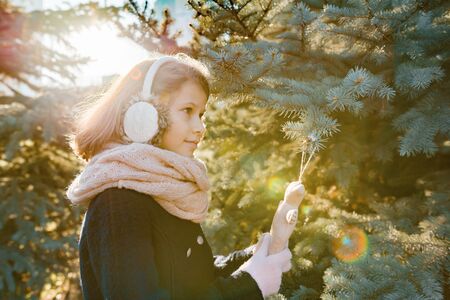 Winter outdoor portrait of child girl near the Christmas tree, smiling girl decorates Christmas tree with toy, golden hour.の写真素材