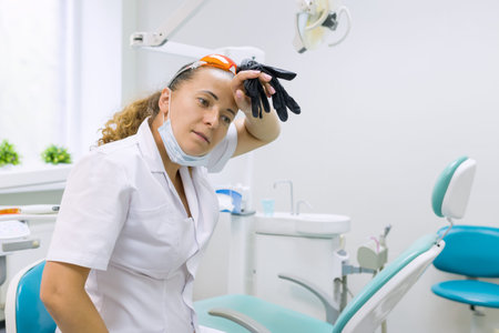 Tired woman dentist, background dental office chair. Female dentist taking off gloves wiping her forehead.の写真素材
