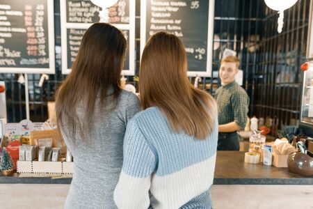 Autumn winter portrait of two young women standing with their backs near the bar counter in coffee shop, girls talking to a barista man.の写真素材