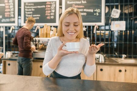 Young smiling barista girl with cup of coffee. Blonde woman in apron enjoying the aroma of fresh coffee, near a bar counter in coffee shop.の写真素材