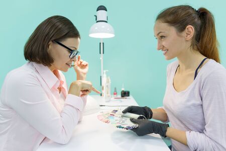 Young beautician girl manicurist talks to a female client, does manicure, chooses the color of nail polish, communicates and smiles.の写真素材