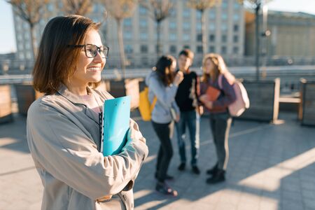 Portrait of mature smiling female teacher in glasses with clipboard, outdoor with a group of teenagers students, golden hour.の写真素材