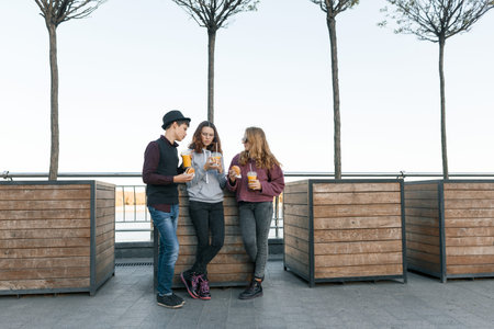 Teenagers eat street food, friends boy and two girls on city street with burgers and orange juice....
