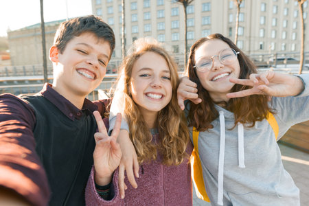 Portrait of three teen friends boy and two girls smiling and taking a selfie outdoors. City background, golden hour.の写真素材