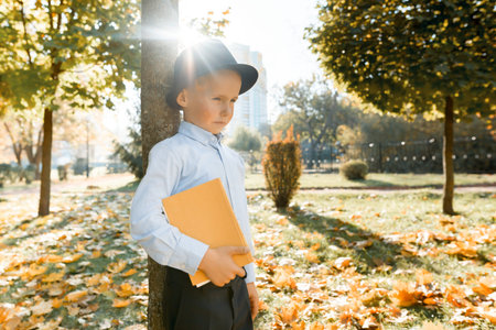 Offended little boy in hat, classic shirt holding a book in his hands. Stands near a tree in sunny...