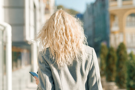 View from the back of young blonde woman walking along city street, autumn sunny day.