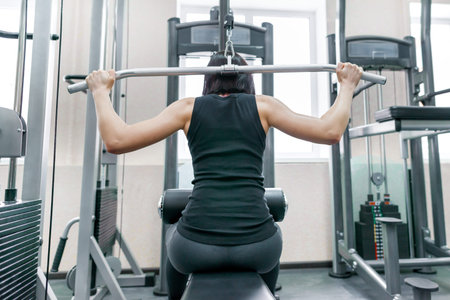 Young woman doing exercises for the back on a fitness machine in the gym, view from the back....