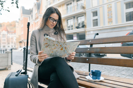 Young beautiful woman sitting on bench with travel suitcase and reading city map, drinks coffee,...