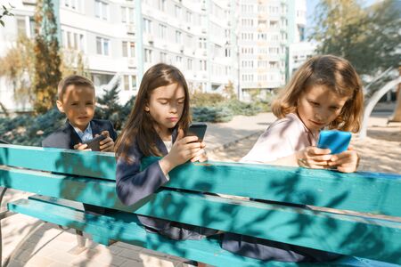 Group of children 7, 8 years old with mobile phones, enthusiastically looking into smartphones, schoolchildren with backpacks, outdoor background. Education, friendship, technology and people concept,の写真素材