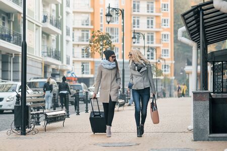 Two young smiling beautiful women in warm clothes walking down the city street with a travel suitcase, women laughing and talking.の写真素材