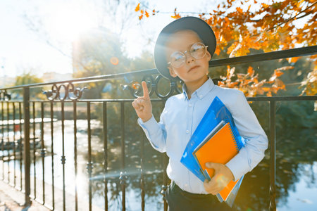 Little smart boy in glasses with books, shows index finger upwards idea, eureka. Background autumn...