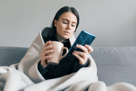 Autumn winter portrait of a young girl resting at home on the sofa with mobile phone and cup of hot drink, under warm blanket.の写真素材