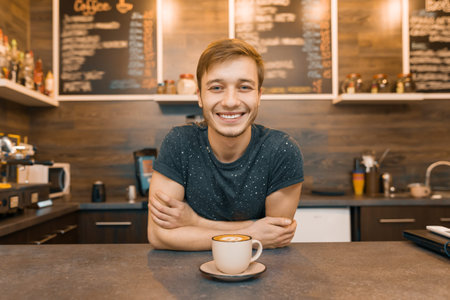 Portrait of young smiling male barista with prepared drink with arms crossed standing behind cafe...