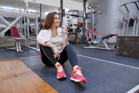 Portrait of smiling mature woman with towel drinking water from bottle in gym. Health fitness sport...
