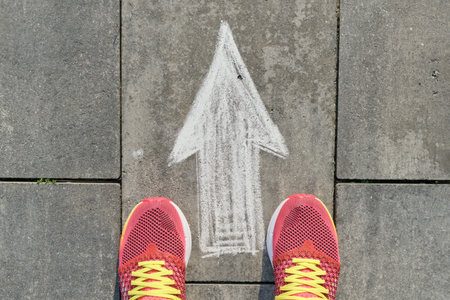 Arrow sign painted on gray sidewalk with women legs in sneakers, top view.