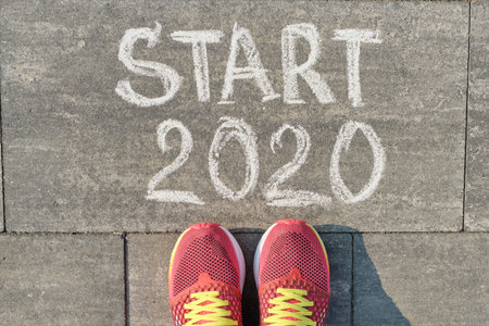 Start 2020, text on gray sidewalk with woman legs in sneakers, top view.
