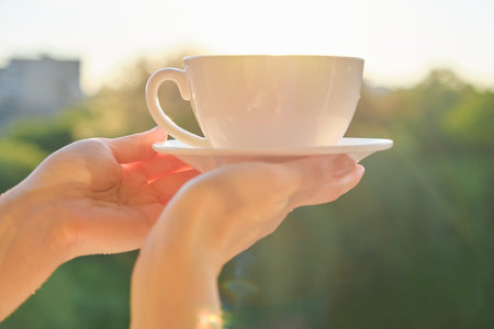 Set of white ceramic cup with saucer in womans hand, background sunset, evening cityの写真素材